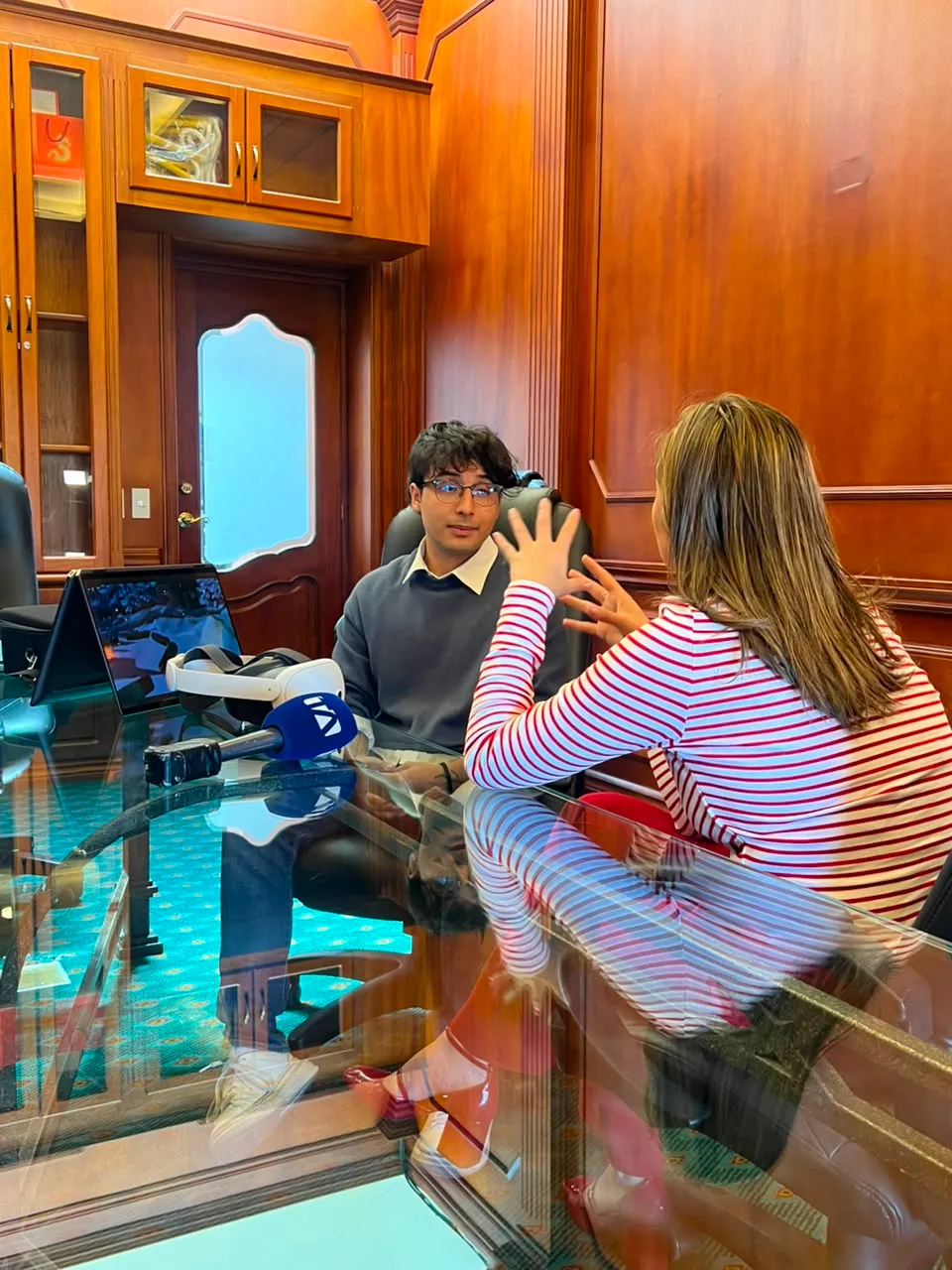 Sebastian being interviewed at a desk by a TV reporter holding a network microphone, with a laptop and a VR headset on the table
